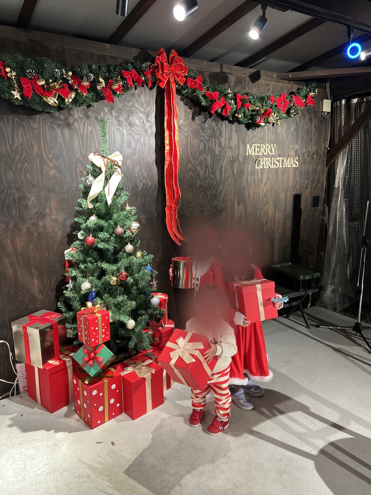 「MERRY CHRISTMAS」の文字とツリー、プレゼントの箱が並ぶフォトスポットでサンタの衣装を着た子どもたち