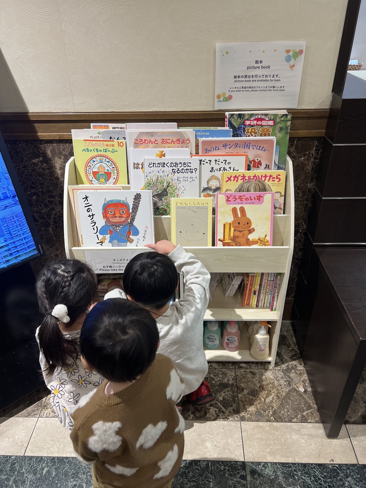 絵本がずらりと並ぶ棚を選ぶ子どもたち。「絵本の貸出を行っております」の案内