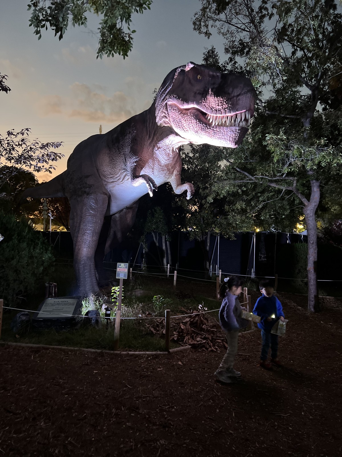 夕暮れの木立に立つティラノサウルスの実物大模型(ナイトツアー時に撮影)