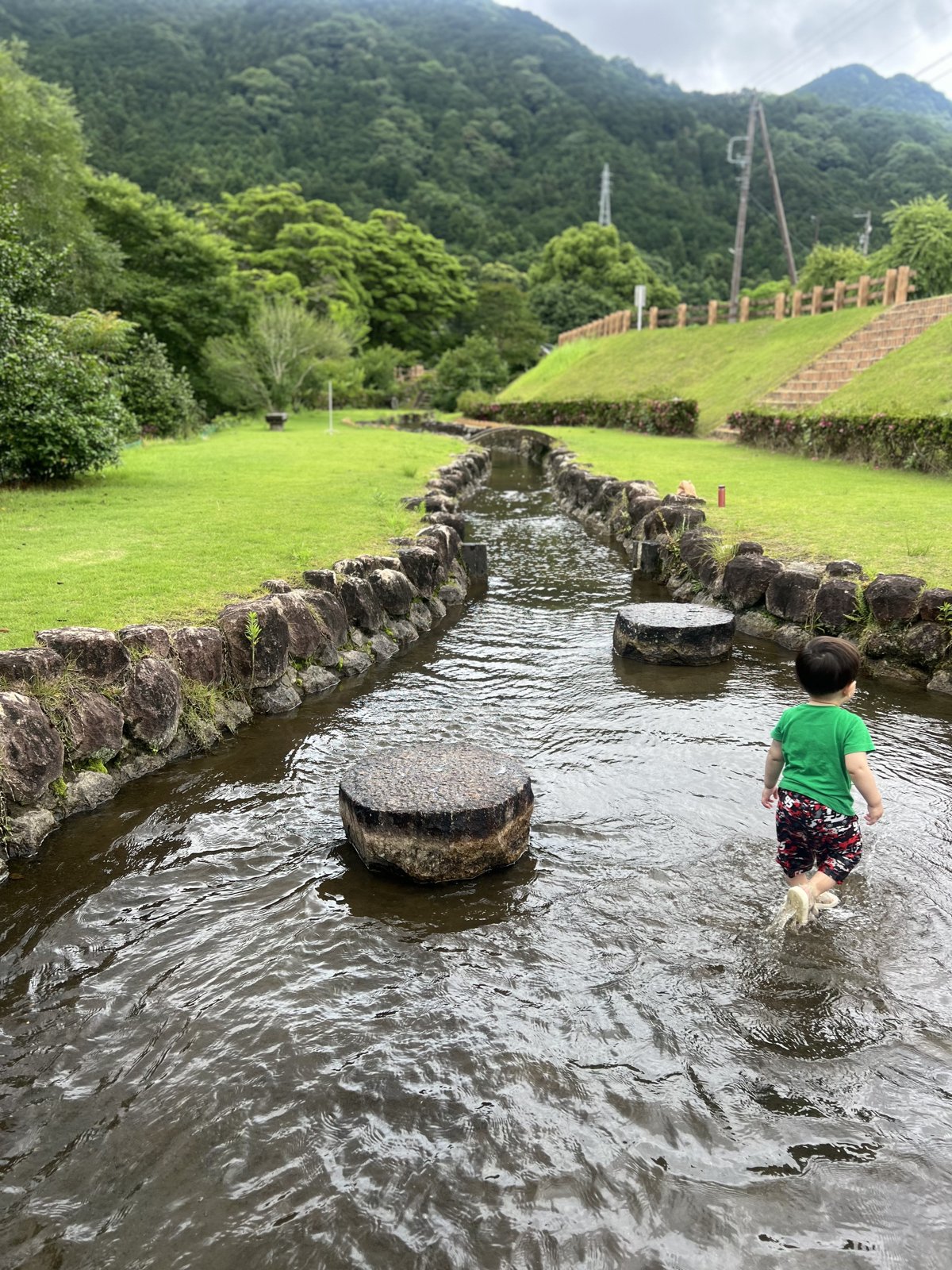 山を背に、石で囲まれた浅い水路を歩く息子