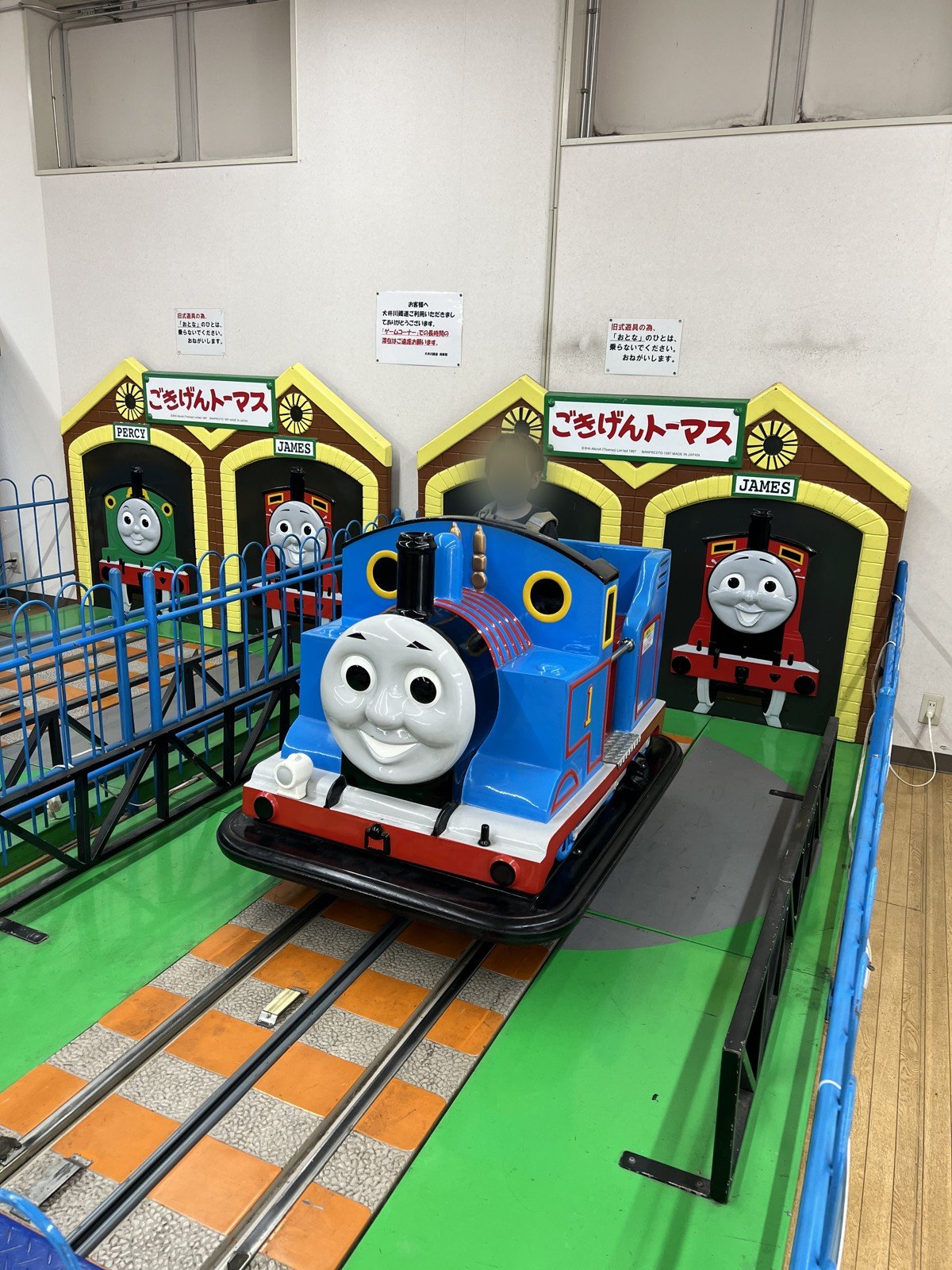 「ごきげんトーマス」の乗り物に乗る次男。うしろにパーシーとジェームスの車庫