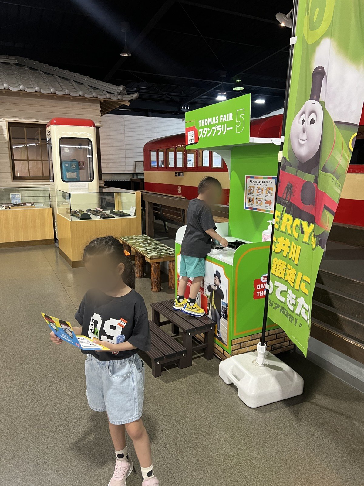 「THOMAS FAIR スタンプラリー」の台でスタンプを押す息子と、台紙を見る娘