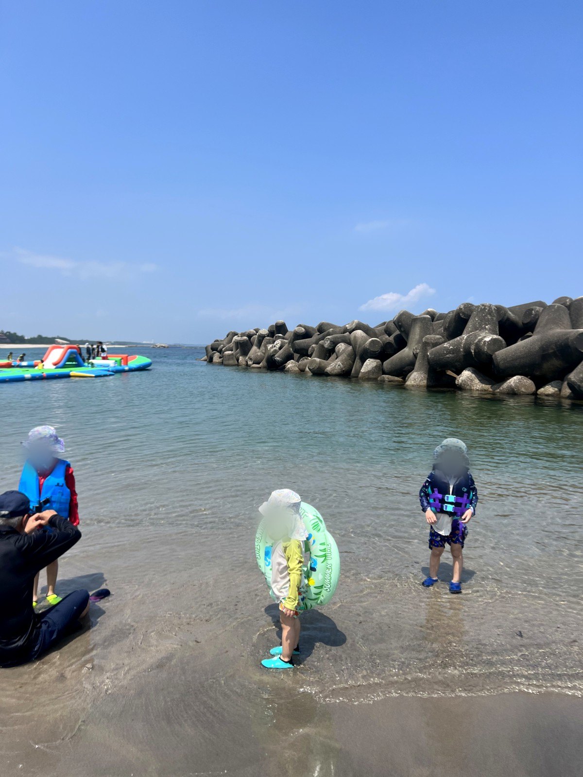 浅瀬でライフジャケットや浮き輪をつけて遊ぶ子どもたち。奥に海上アスレチック