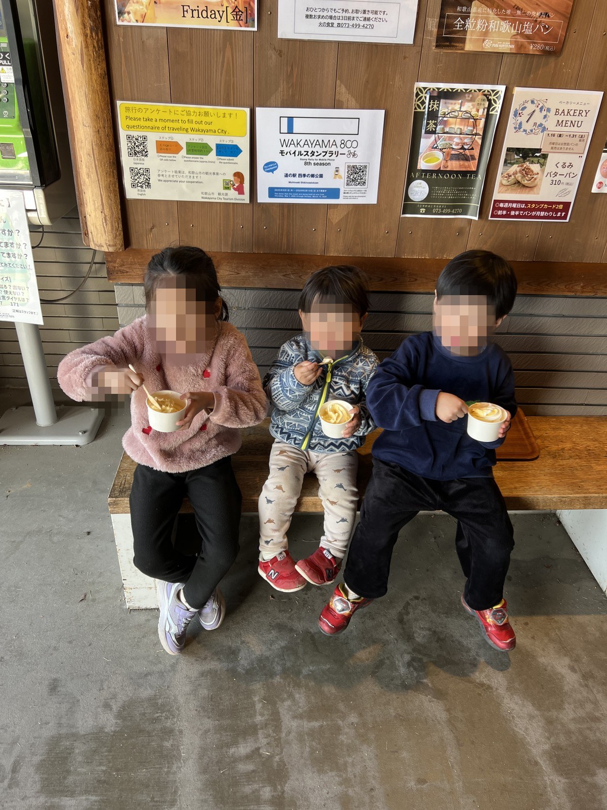 ベンチに並んで座り、カップのソフトクリームを食べる子ども3人